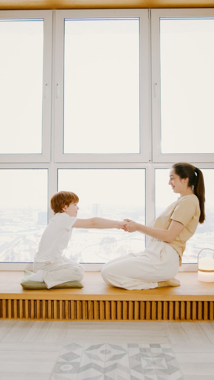 A mother and son lovingly hold hands while sitting by a large window indoors.
